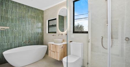 Modern bathroom with green tiled accent wall, freestanding bathtub, floating vanity with round mirror, glass shower enclosure, and large window.