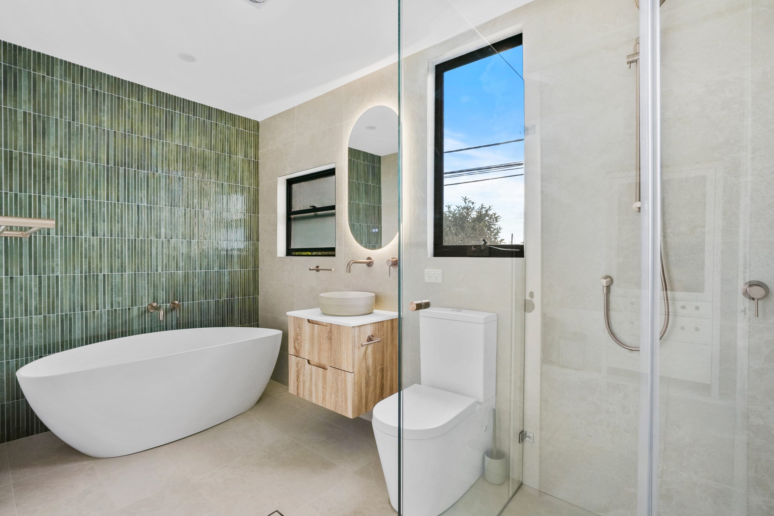 Modern bathroom with green tiled accent wall, freestanding bathtub, floating vanity with round mirror, glass shower enclosure, and large window.