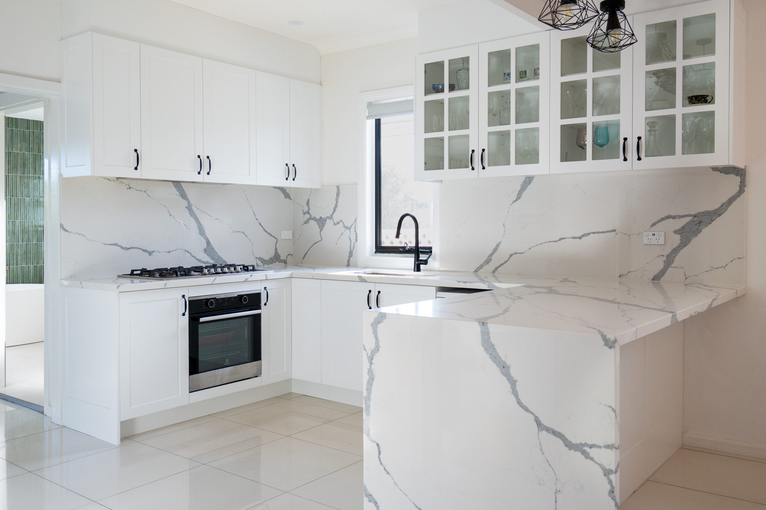 Modern white kitchen with marble-look countertops and backsplash, built-in oven, gas cooktop, and glass-front upper cabinets.