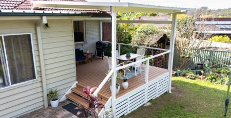 Covered outdoor deck with white railing, wooden flooring, and outdoor furniture including a blue sofa, white chairs, and dining table.