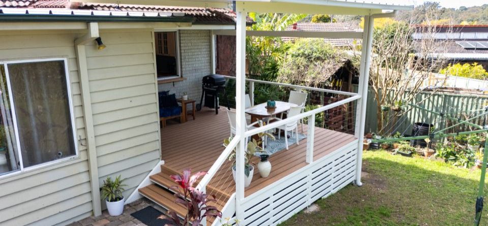 Covered outdoor deck with white railing, wooden flooring, and outdoor furniture including a blue sofa, white chairs, and dining table.