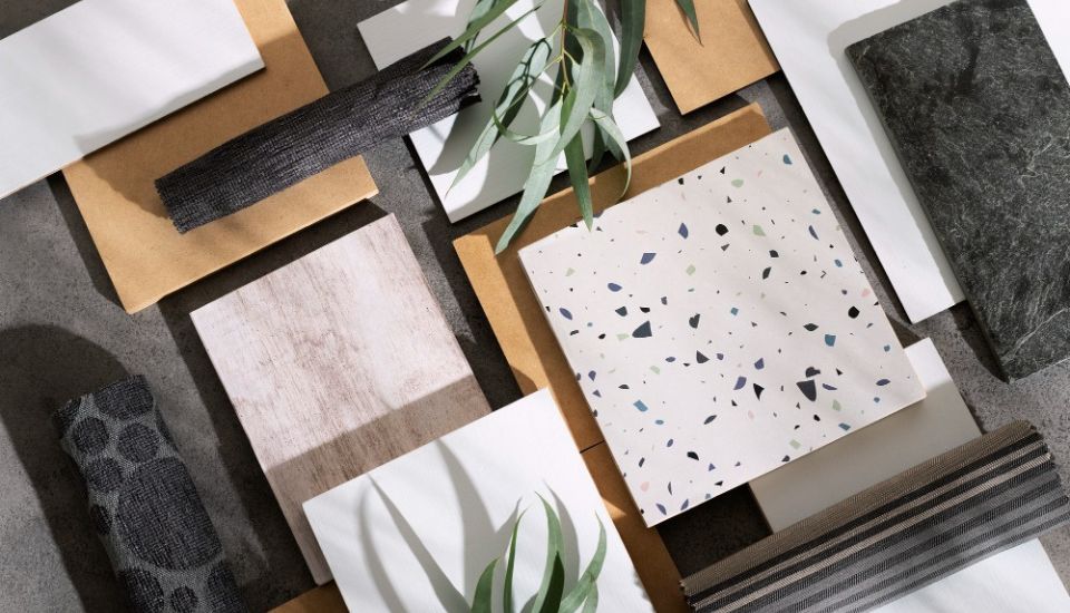 Flat lay of modern renovation materials including terrazzo tile, wood samples, stone, and fabric swatches arranged aesthetically on a grey surface with green leaves.
