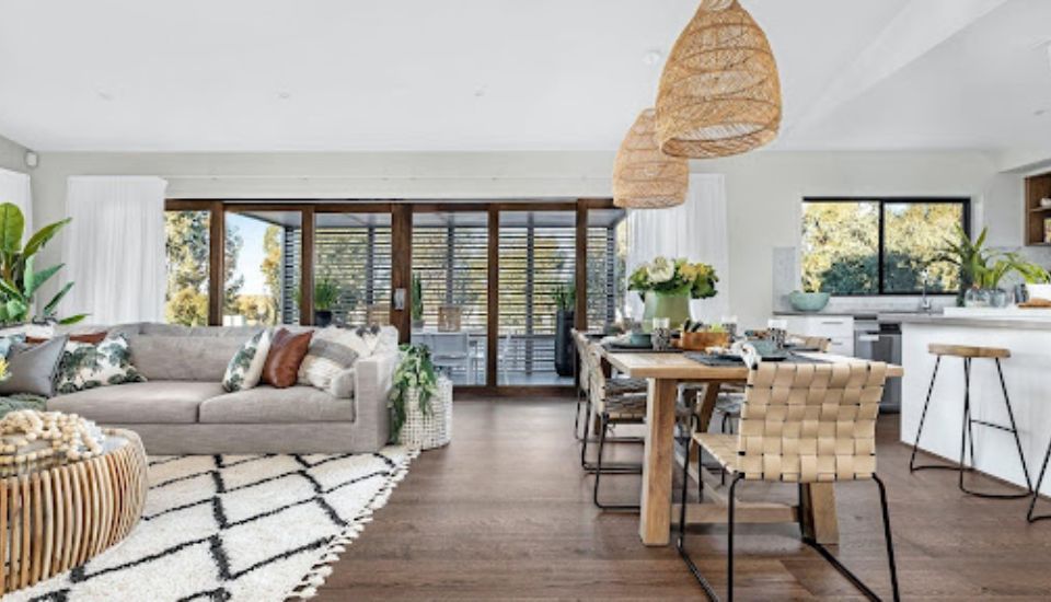 Modern open-plan living and dining area with wooden flooring, woven pendant lights, neutral furniture, and large glass doors leading to an outdoor space.