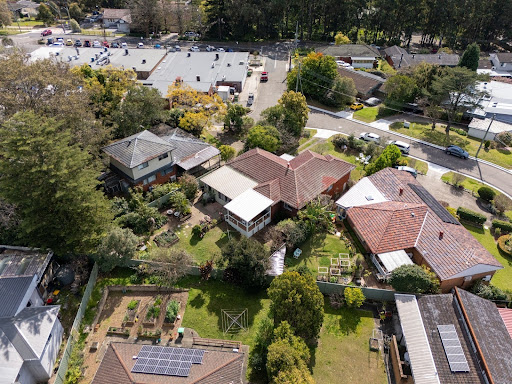 Aerial view of suburban houses with gardens, backyards, and a nearby parking lot surrounded by trees.
