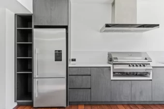 Outdoor kitchen with stainless steel fridge, built-in barbecue grill, and modern grey cabinetry.